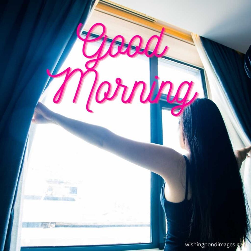 A young black-haired girl opens the blue colored curtains and looks outside through the window in the morning