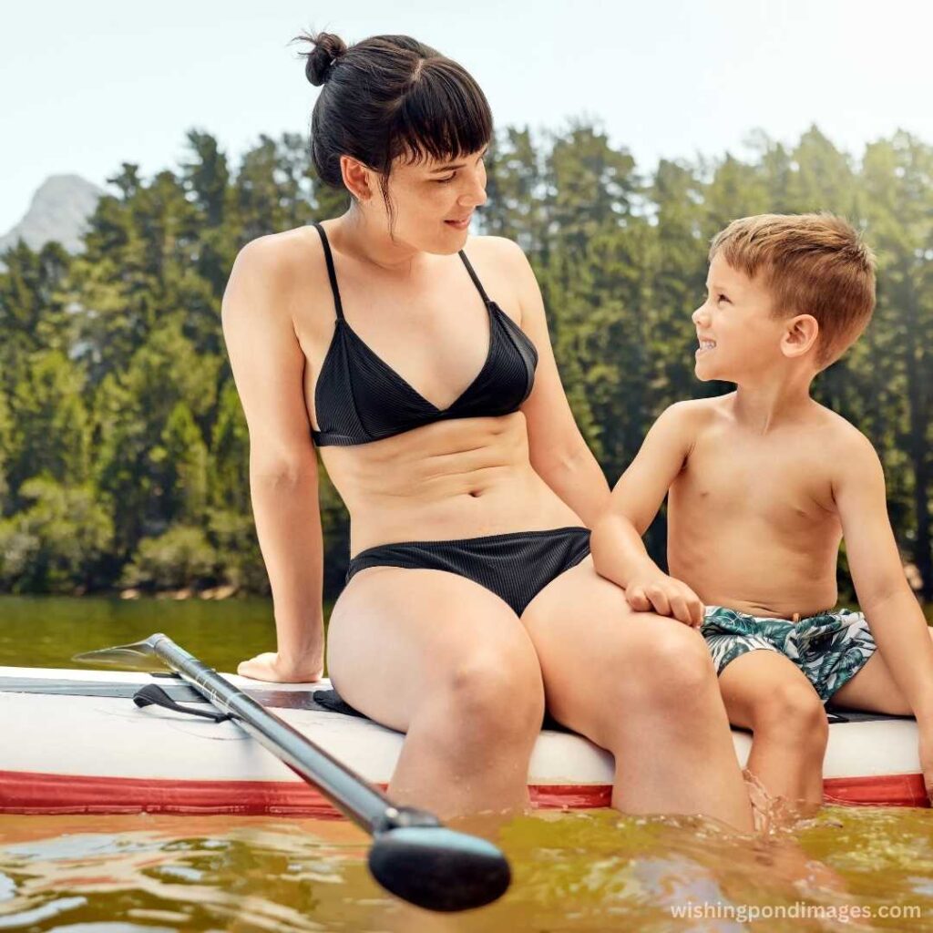 Woman wearing the bikini costume sitting in the paddle board with her son in the lake near mountain trees - Nature Images
