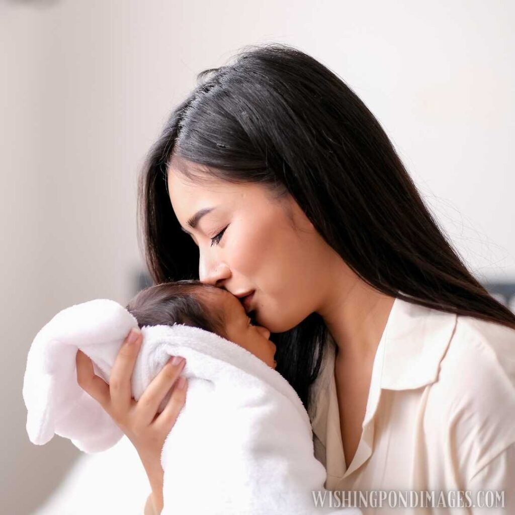 White shirt Asian mother is kissing at forehead of her newborn baby in bedroom in front of glass windows. Newborn baby image