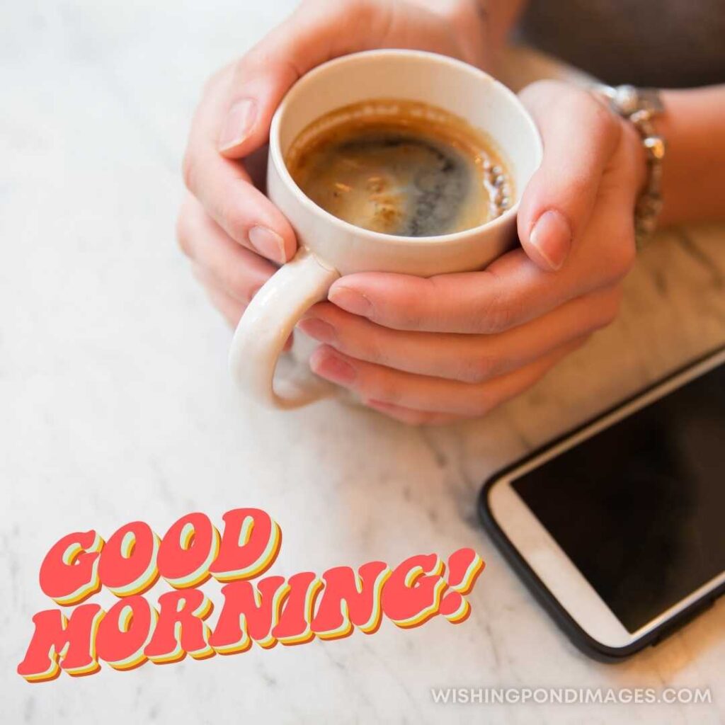 A girl holding a cup of coffee in her hands with phone sitting on the table. Good Morning Coffee Images
