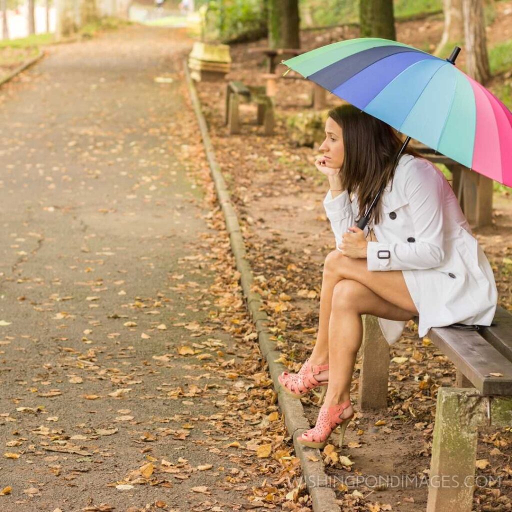 Beautiful angry girl sitting alone and waiting under a rainbow umbrella. Feeling alone images girl.