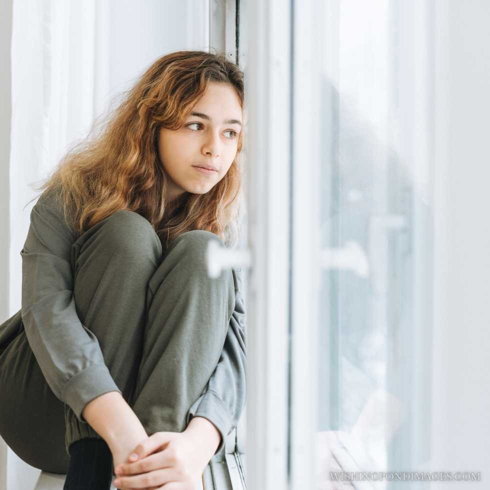 Beautiful sad unhappy teenage girl with curly hair sitting on the window sill. Sad girl in room.