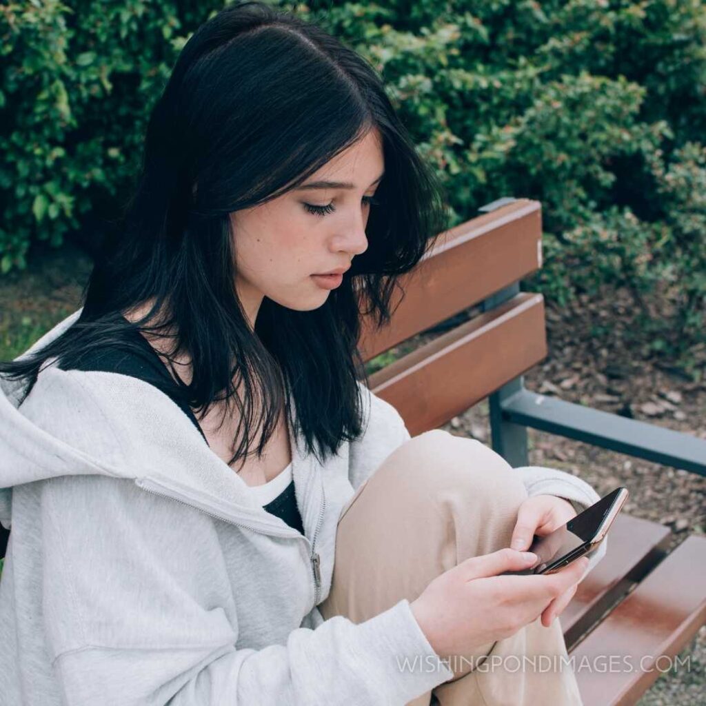 Beautiful teenage girl using smartphone while sitting alone on the bench outdoors on the street. Feeling alone images girl.