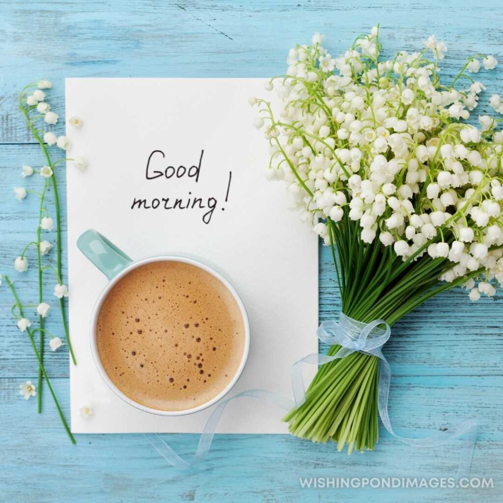 Good Morning note on the paper with a cup of coffee with white flowers on the light blue-colored table. Good Morning Coffee Images