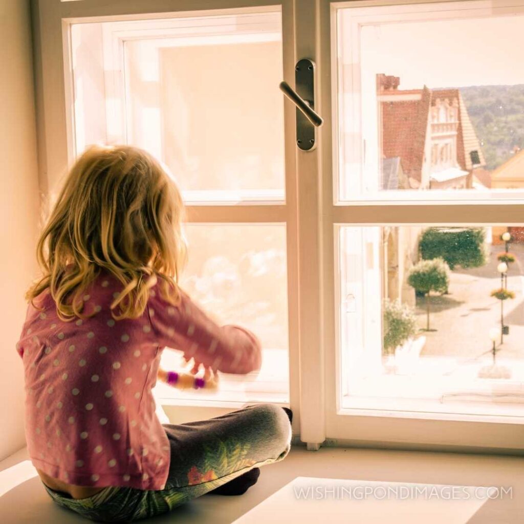A little blonde girl sitting and looking over the window. Feeling alone images girl.