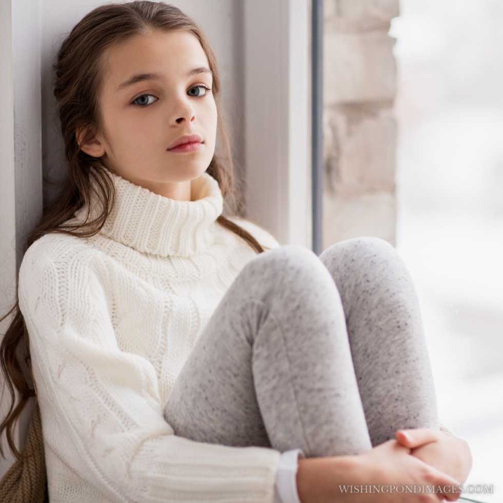 Sad beautiful girl in a sweater sitting on the sill at the home window in winter. Sad girl in room.