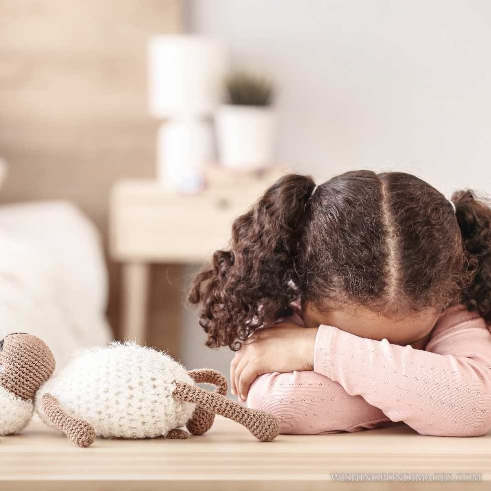 Sad little African-American girl sitting at the table. Sad girl in room.