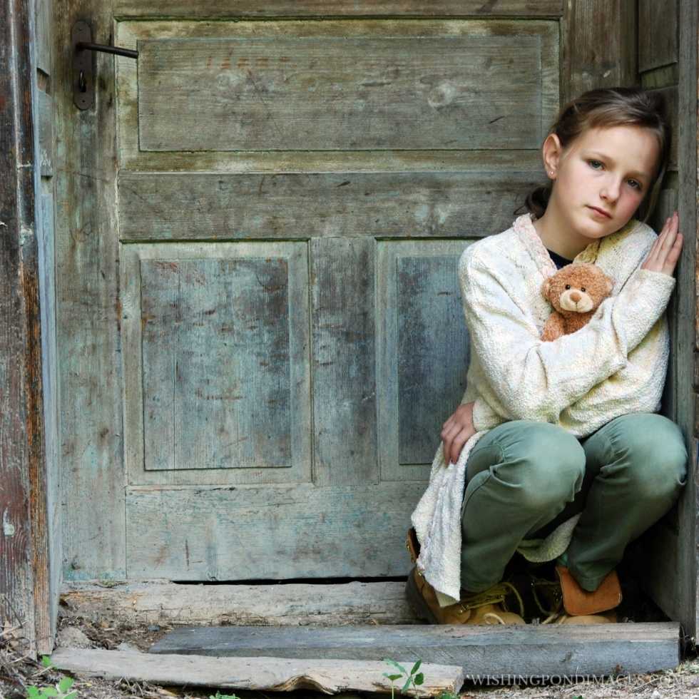 Sad little girl leaning on an old wooden door outdoors with a stuffed teddy bear. Sad girl in room.