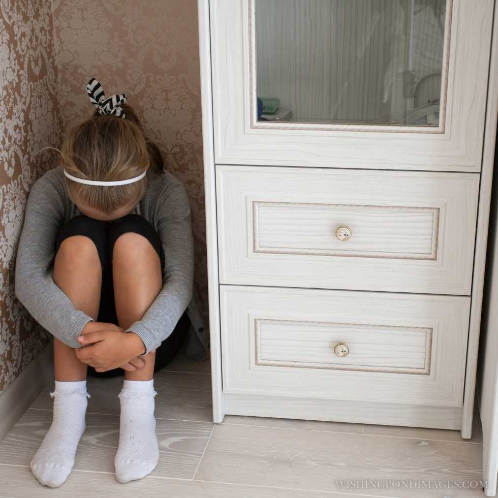 A sad little girl sitting in the corner of a room head lowered, legs clasped by her hands. Sad girl in room.