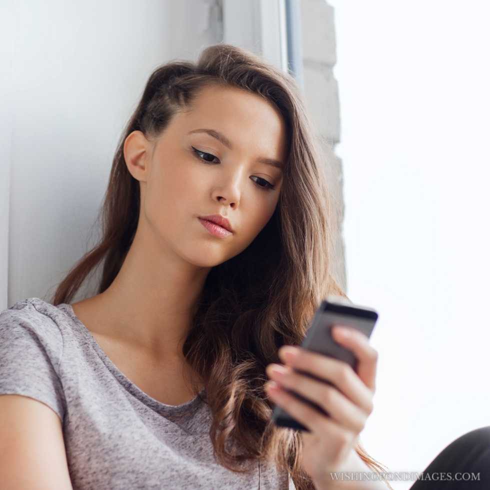 Sad unhappy pretty teenage girl sitting on the windowsill with a smartphone and texting. Sad girl in room.