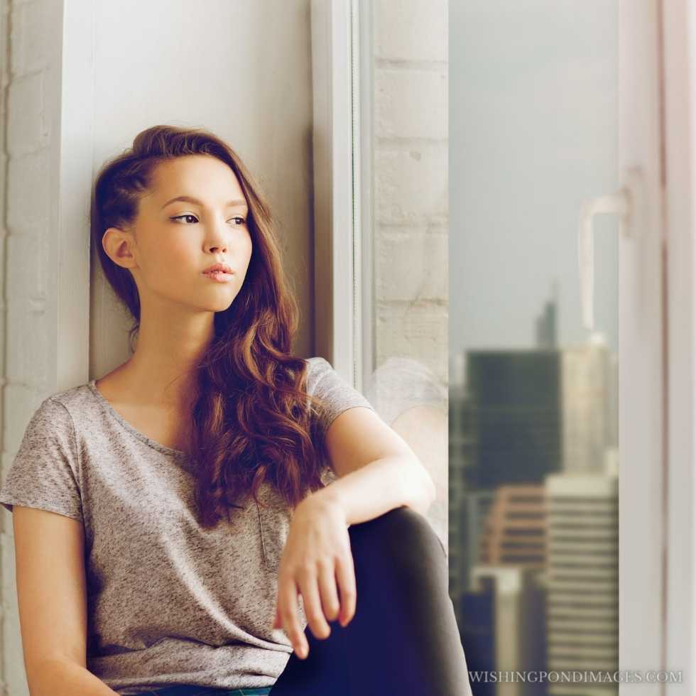 Sad unhappy pretty teenage girl sitting on the windowsill. Sad girl in room.