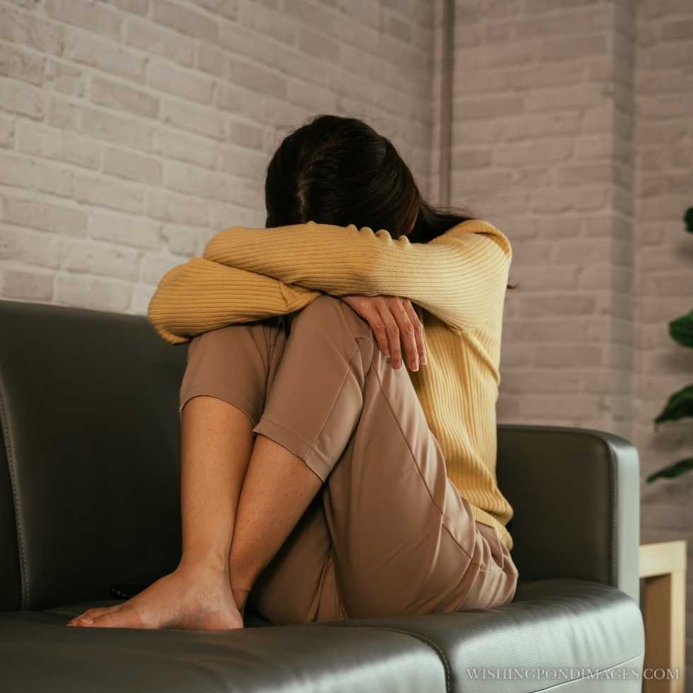 A young woman sitting on the couch at home hiding her face and hugging her knees while feeling depressed. Sad girl in room.